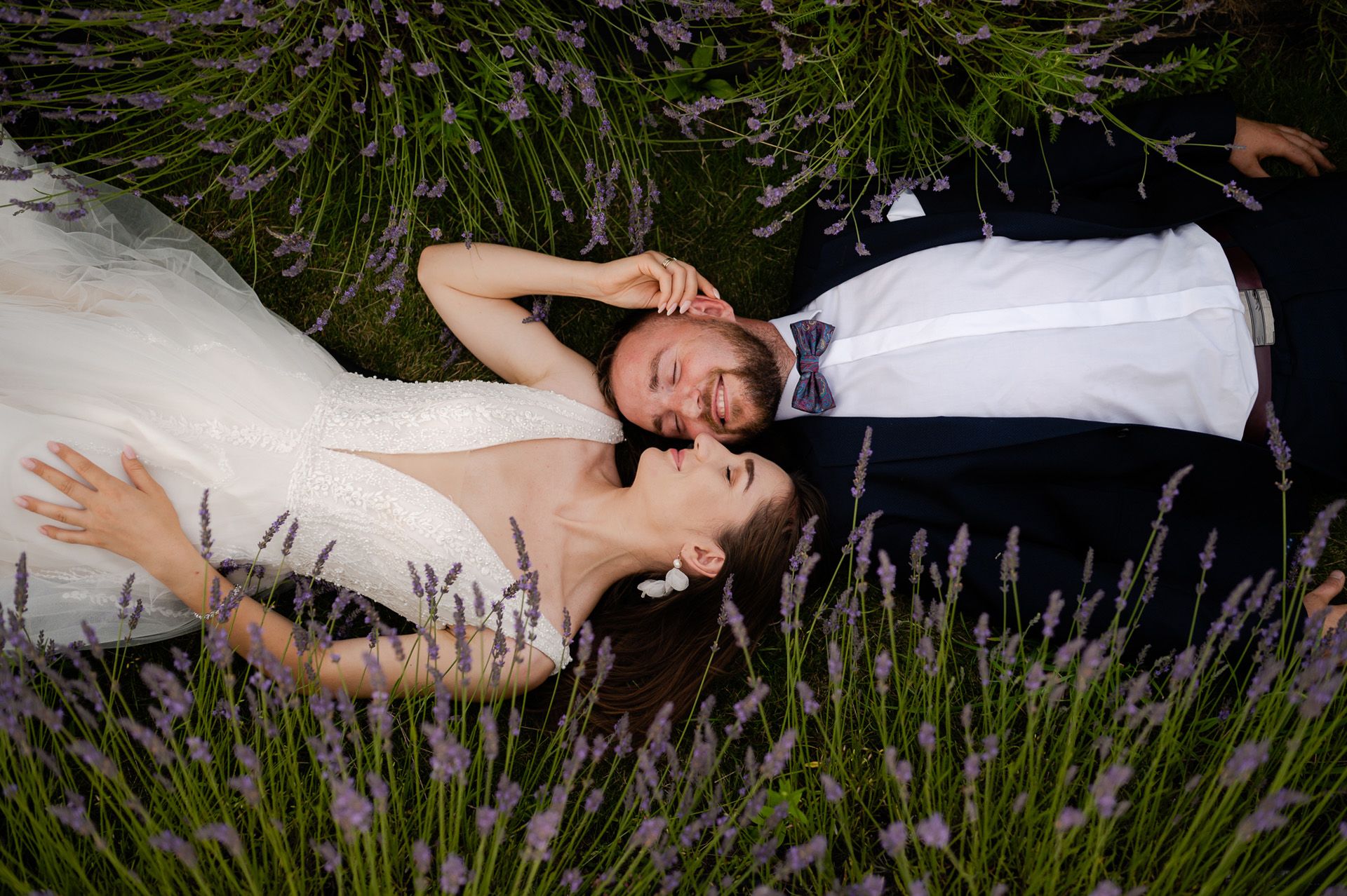 Para młoda leży w polu lawendy podczas sesji ślubnej - panna młoda w białej sukni i pan młody w granatowej kamizelce z muszką relaksują się wśród fioletowych kwiatów, romantyczna sesja plenerowa