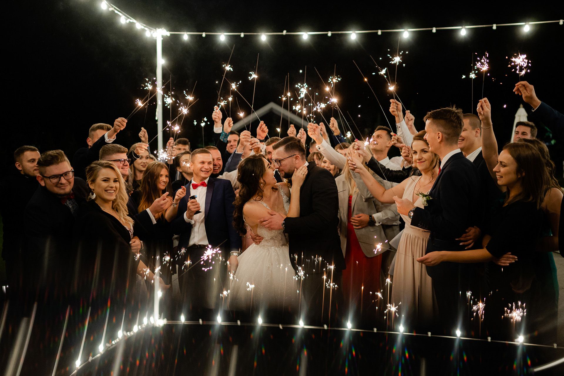 Para młoda całuje się otoczona gośćmi trzymającymi zimne ognie, romantyczny moment ślubu na zewnątrz z lampkami w tle